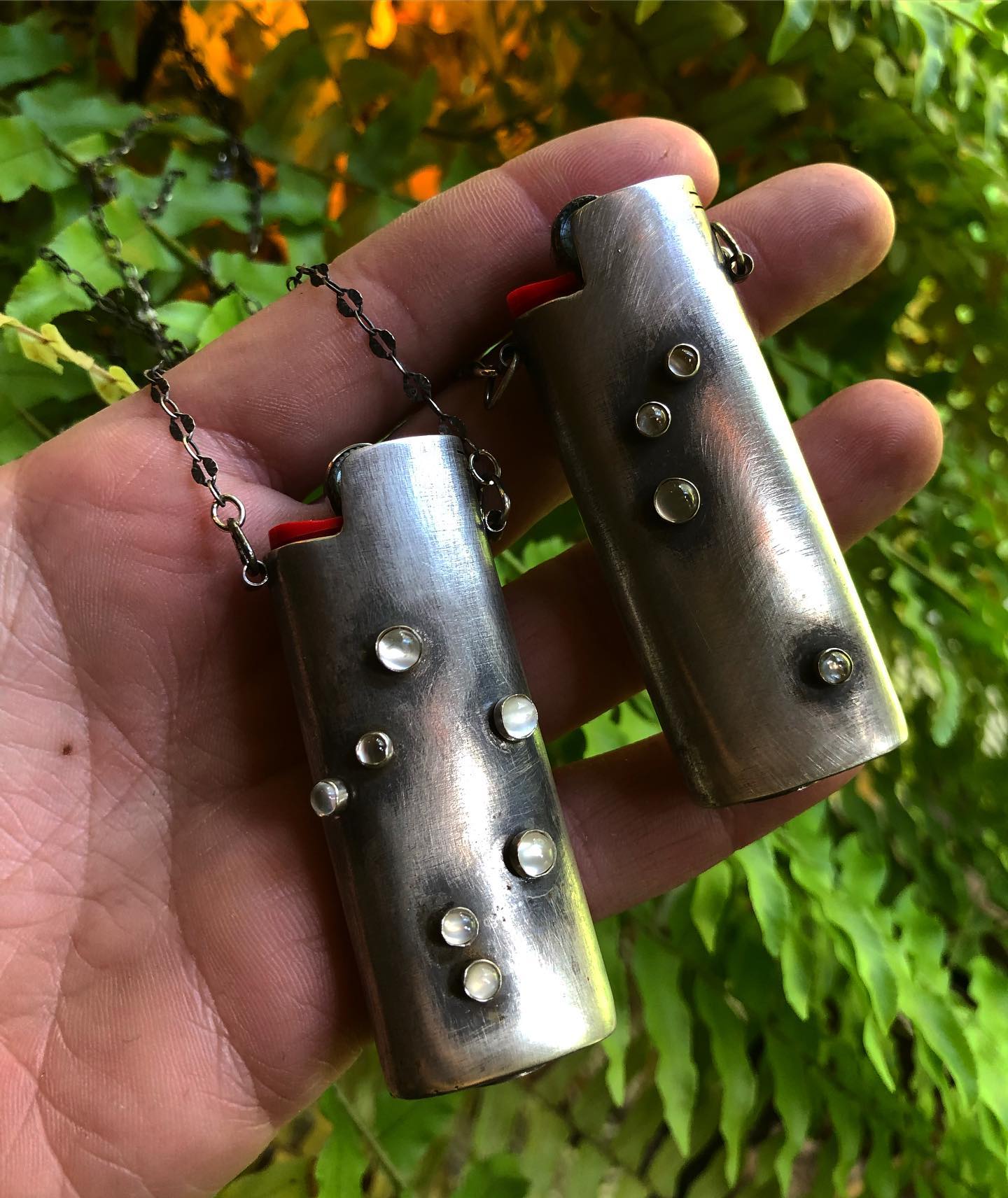 Two sterling Zodiac FireKeeper lighter cases held in a hand against ferns
