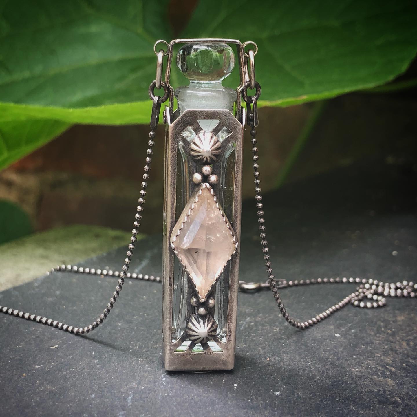 A sterling Quartz PotionKeeper phial with a pink faceted stone, resting on slate beside a large green leaf