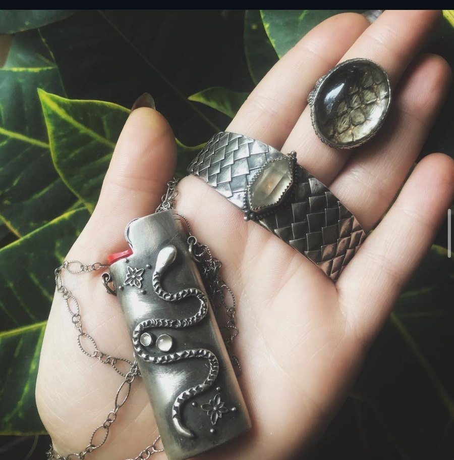 A hand holding a sterling serpent FireKeeper lighter case, wearing a woven scale cuff and a large oval ring, over green leaves